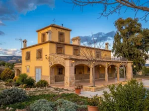 Casa rural de fachada ocre con porche, torre y jardín en un entorno montañoso